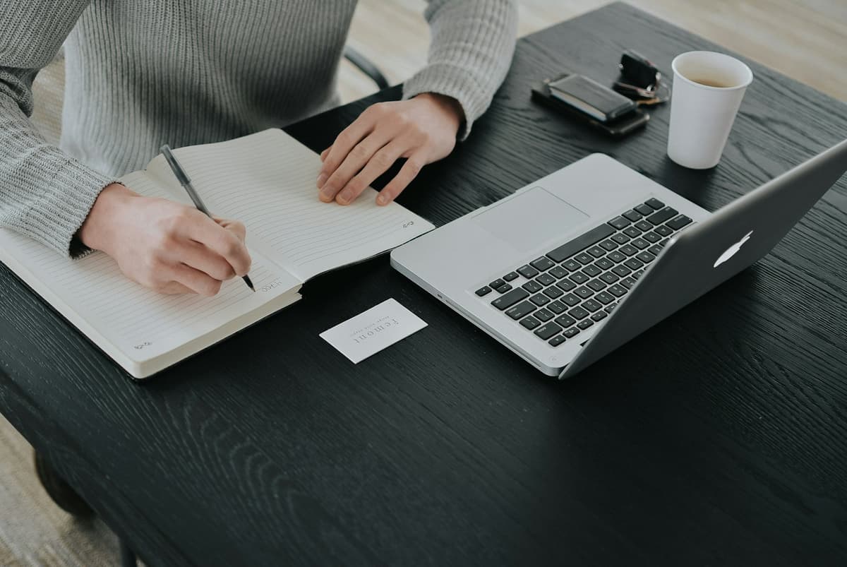Une main écrit dans un carnet à côté d'un ordinateur portable.